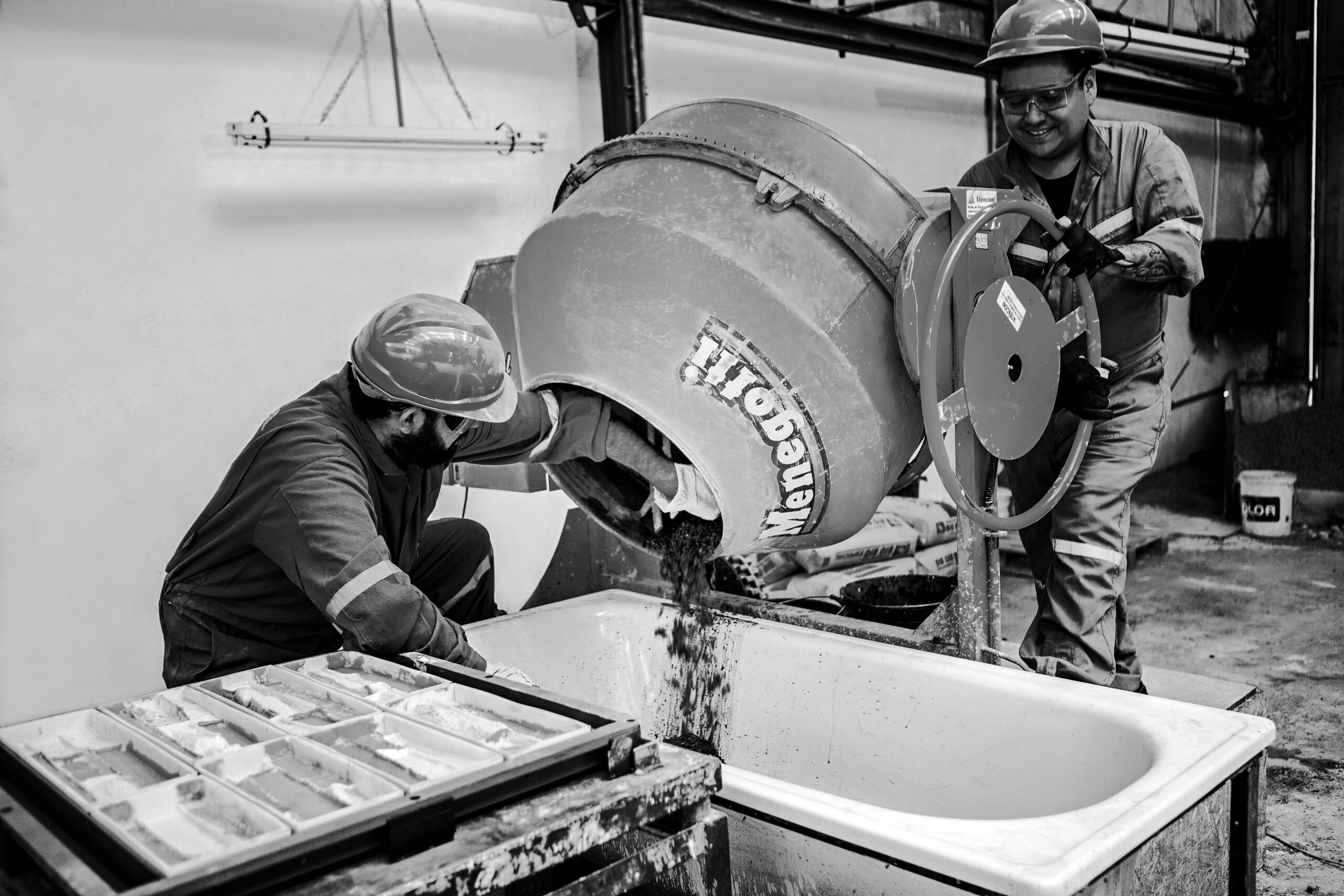 Fotografía en blanco y negro que muestra a dos operarios con cascos de seguridad utilizando una hormigonera industrial para verter la mezcla de cemento y arena en un contenedor, con moldes de producción en primer plano.