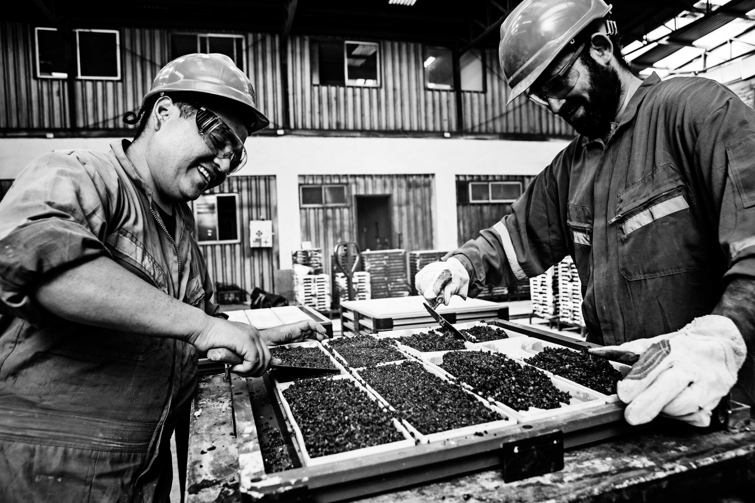 Fotografía en blanco y negro de dos operarios con equipo de seguridad (cascos y gafas) llenando cuidadosamente moldes de múltiples cavidades con una mezcla de concreto, utilizando espátulas para nivelar el material.